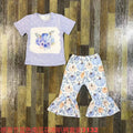 A children's two-piece clothing set featuring a light purple short-sleeve t-shirt with a blue watercolor pumpkin graphic and white speckles, paired with matching flared pants in a blue, purple, and orange pumpkin print. The set is laid flat on a dark wood plank background.