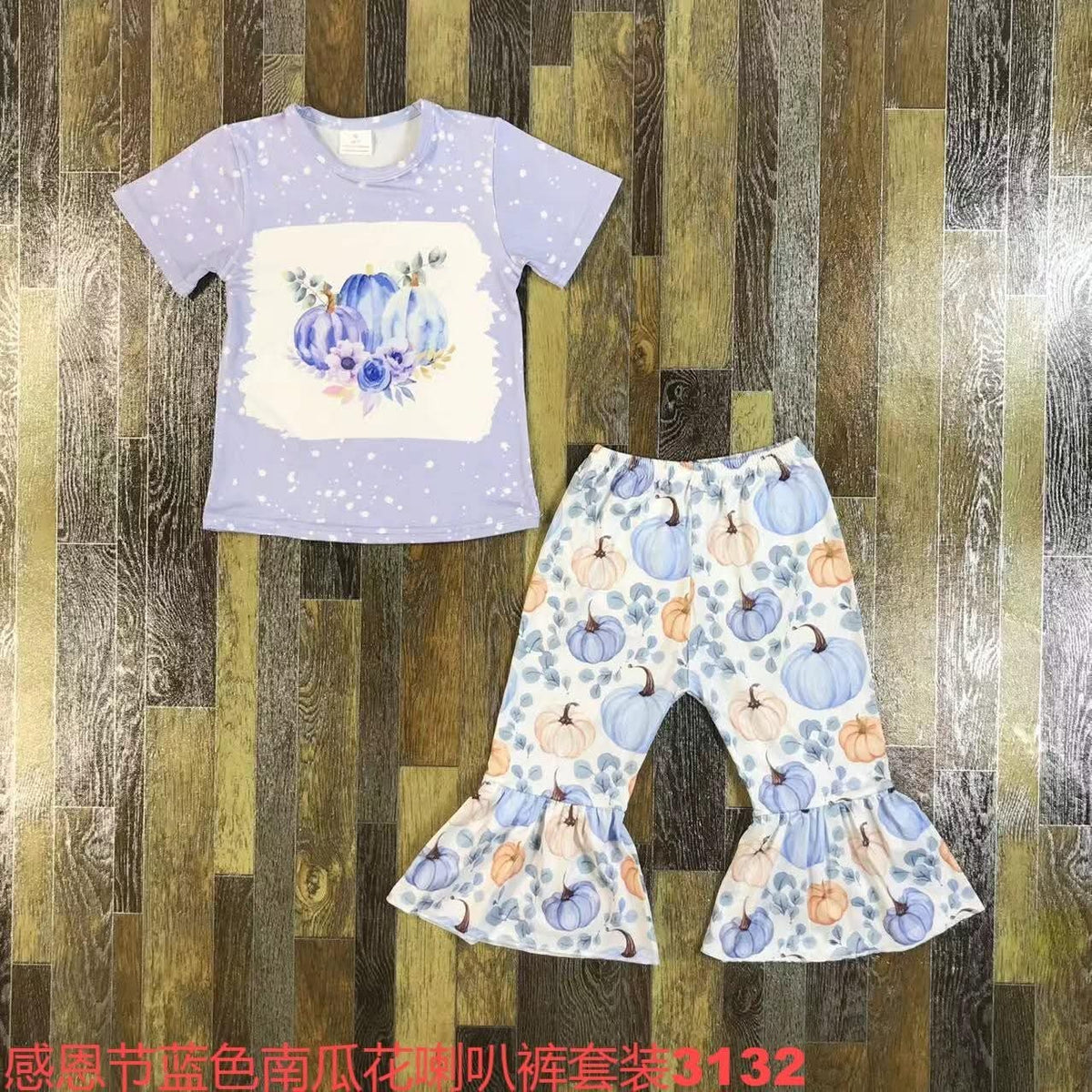 A children's two-piece clothing set featuring a light purple short-sleeve t-shirt with a blue watercolor pumpkin graphic and white speckles, paired with matching flared pants in a blue, purple, and orange pumpkin print. The set is laid flat on a dark wood plank background.